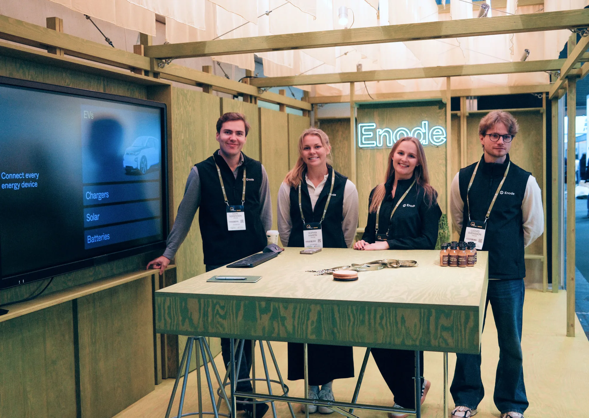 Four Enode team members behind the stand’s timber counter, the neon Enode sign glowing on the wall behind them